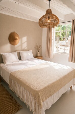 Quarto com cama grande, luminária pendente de fibra natural, um painel redondo de fibra natural na parede, um vaso com flores secas e vista para o verde lá fora.