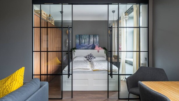 A modern bedroom with a sliding glass door, a bed with a forest-themed painting, a small table, and a chair. - Sunny 1BR Apartment near Park & Stadium (Riga)