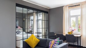 A modern living room with a grey sofa, a yellow cushion, and a colorful pillow. There is a glass partition with a bed visible behind it, and a dining area with chairs and a table. - Sunny 1BR Apartment near Park & Stadium (Riga)