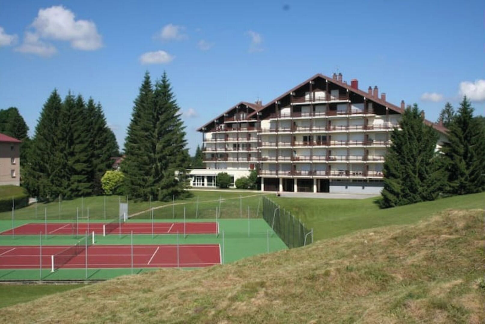 Un hôtel à plusieurs étages, doté de balcons, entouré d’un court de tennis et d’arbres.