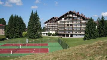 Un hôtel à plusieurs étages, doté de balcons, entouré d’un court de tennis et d’arbres.