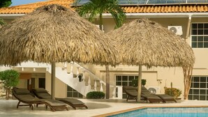 A pool area with two thatched umbrellas, lounge chairs, and a staircase leading to a building with a tiled roof. - 3 Persons - Apartment (Willemstad)