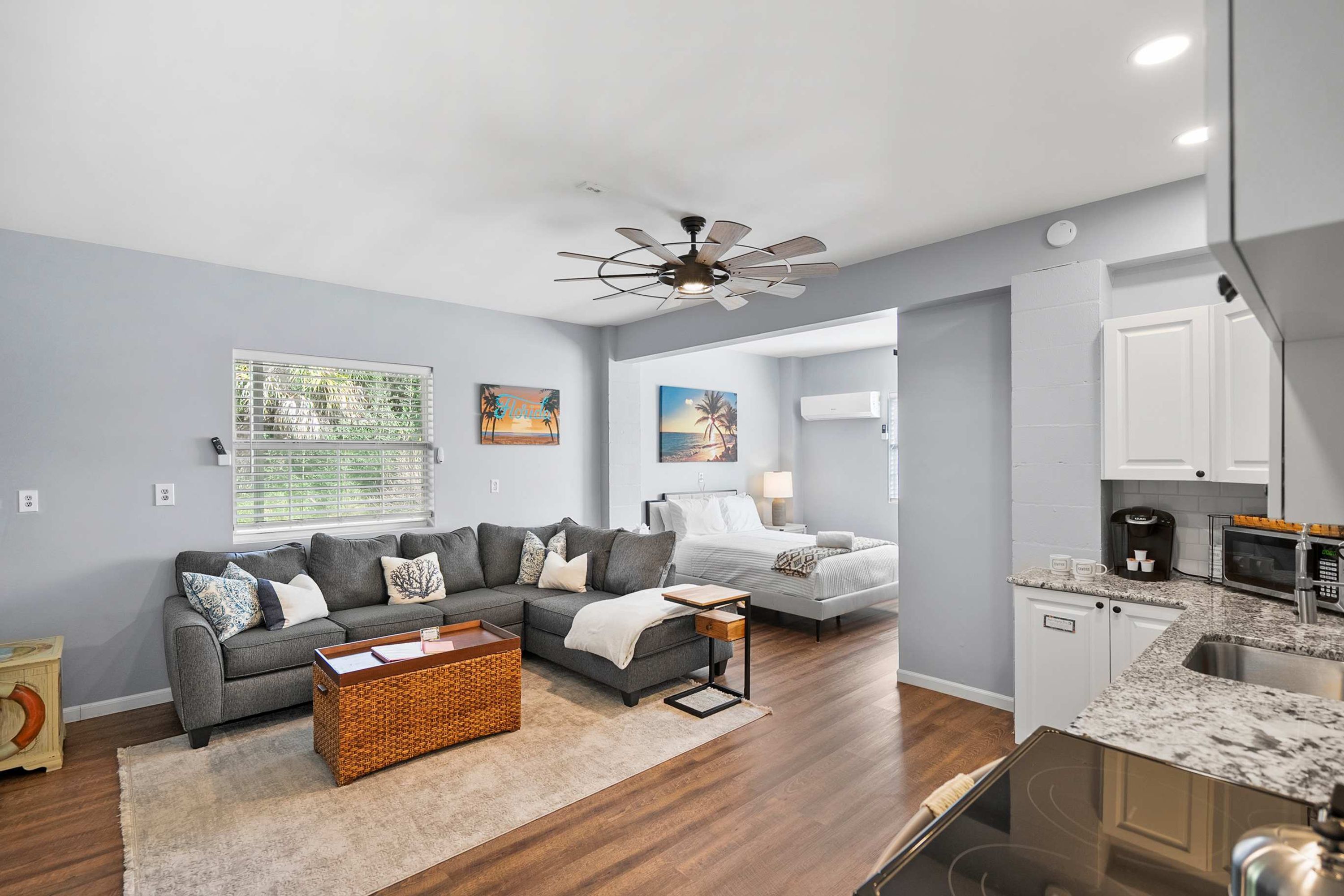 A modern living room with a grey sofa, a coffee table, and a TV. There is a kitchen area with a microwave and a sink.