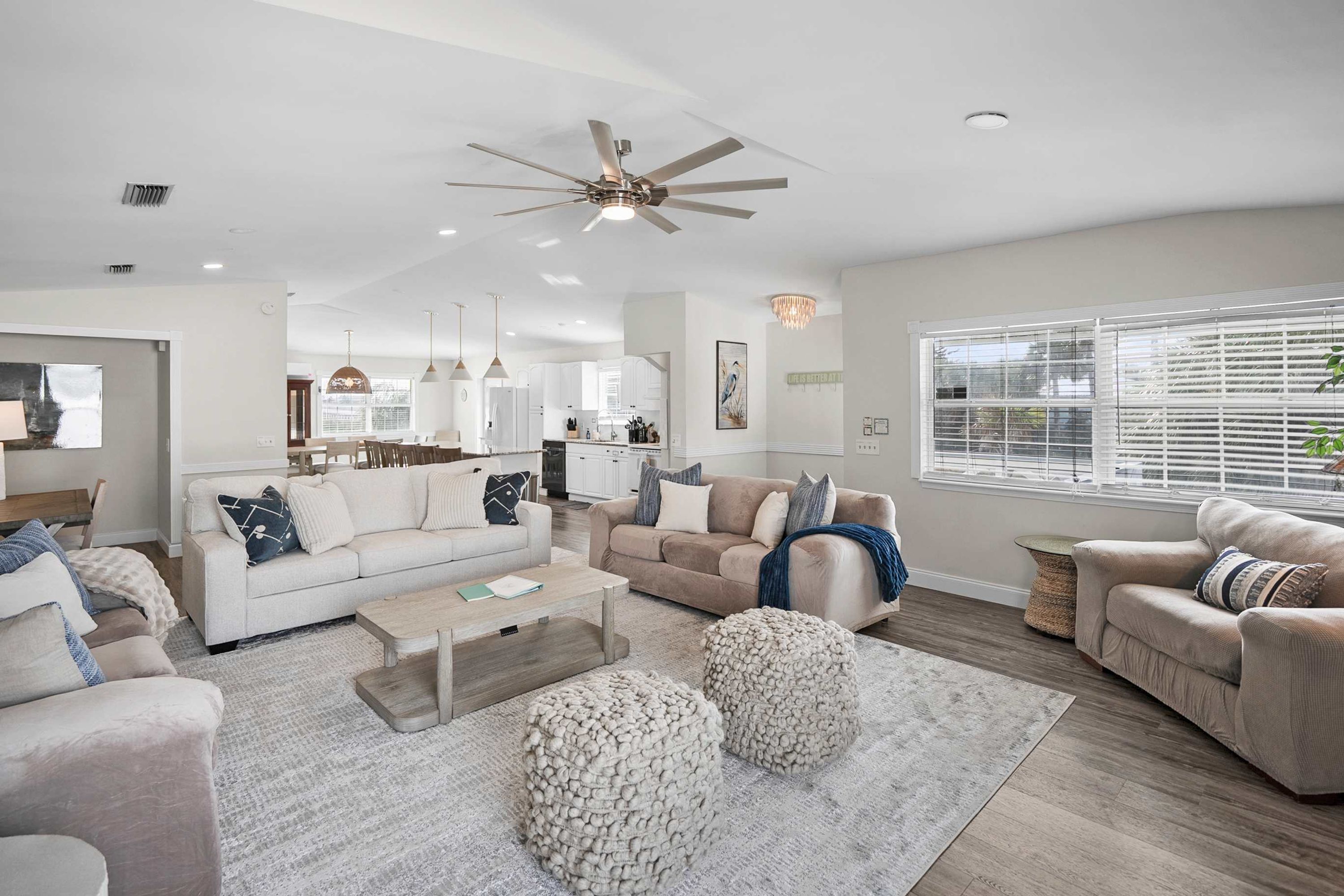 A spacious living room with a high ceiling fan, a large window with blinds, and a kitchen in the background.
