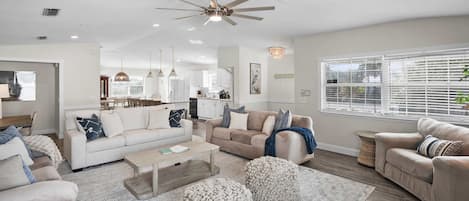 A spacious living room with a high ceiling fan, a large window with blinds, and a kitchen in the background.