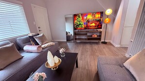 Una sala de estar moderna con un sofá gris, una mesa de centro negra con objetos decorativos, un televisor que muestra una escena navideña y una lámpara de pie.