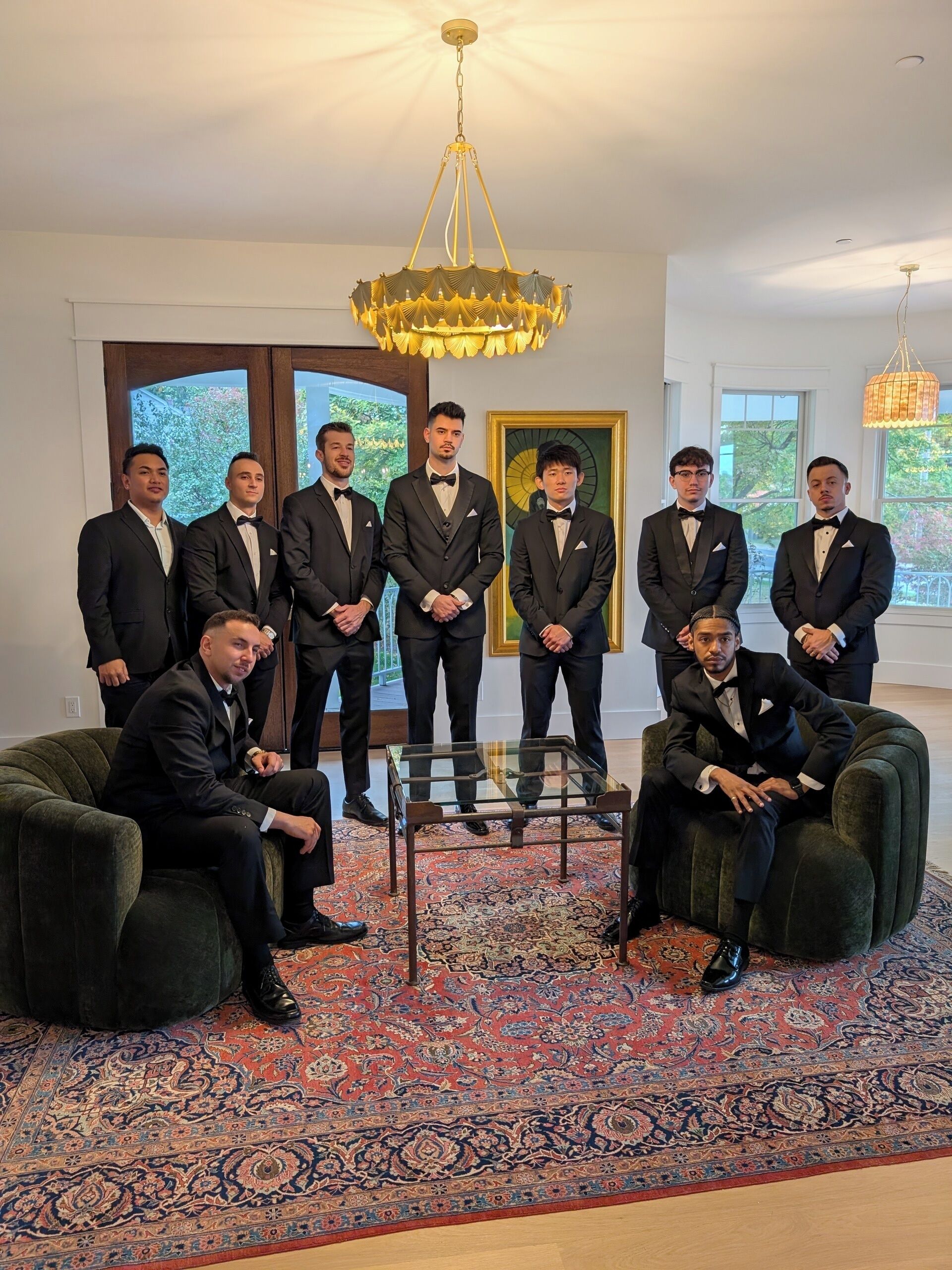 Un groupe d’hommes, vêtus de tenues élégantes, se tient debout et est assis dans une pièce bien éclairée, dotée de grandes fenêtres, d’un lustre et d’un tapis à motifs.