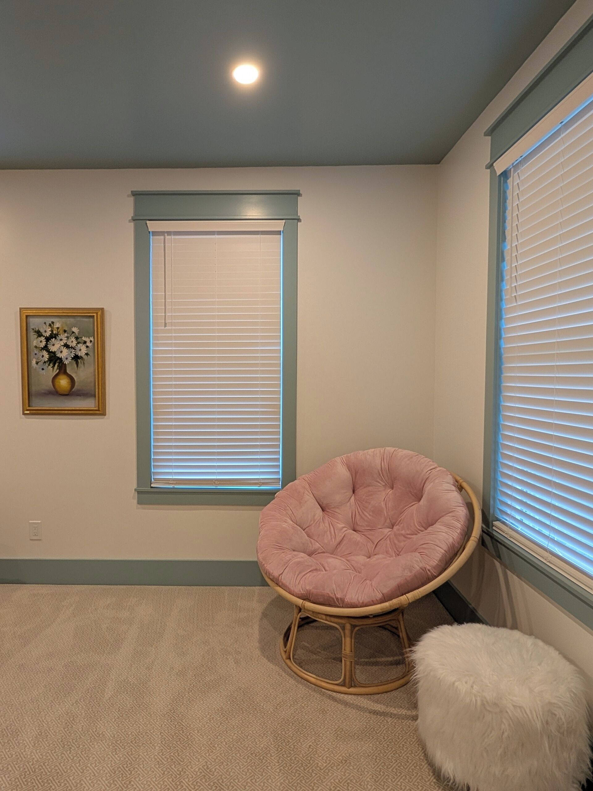 A room with a large window, a chair, a small stool, and a framed picture on the wall.