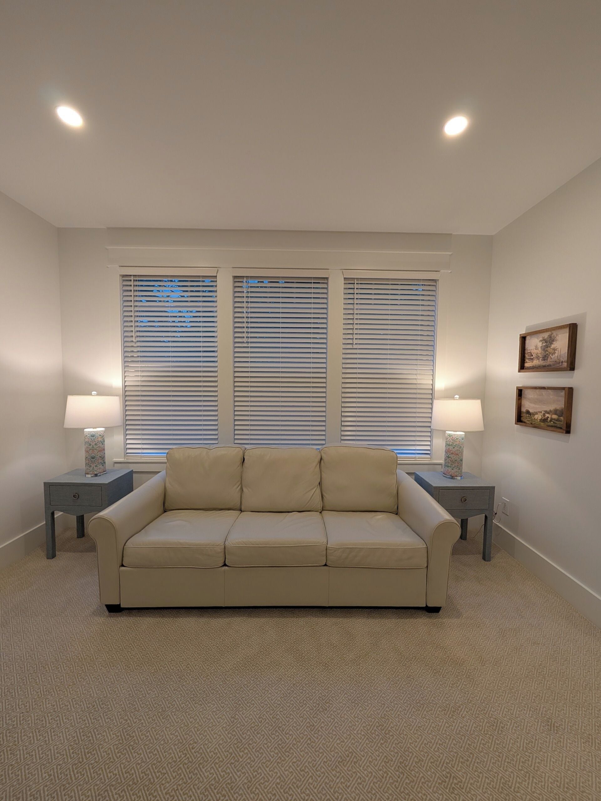 A living room with a beige sofa, two side tables with lamps, and three windows with blinds.