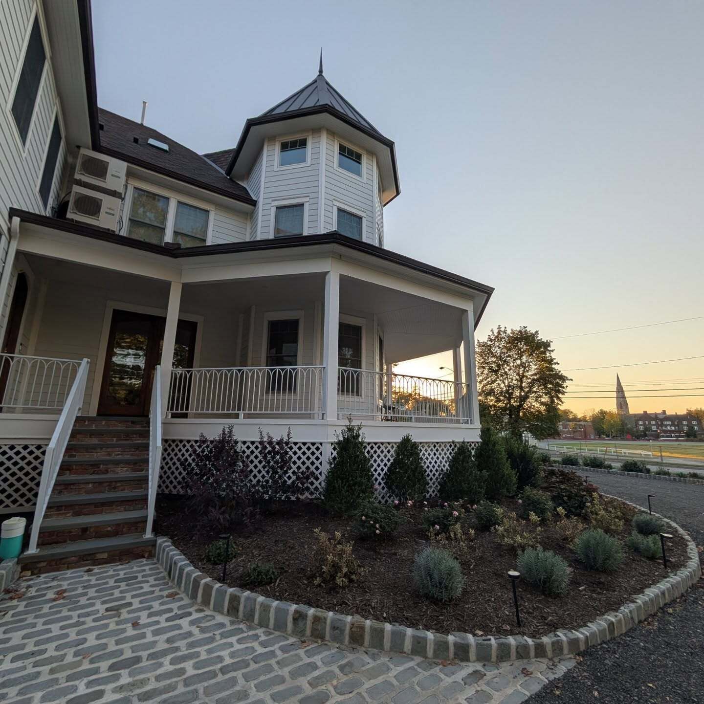 A two-story house with a wrap-around porch, a small tower-like structure on the second floor, and a landscaped garden area.
