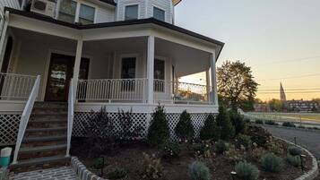 A two-story house with a wrap-around porch, a small tower-like structure on the second floor, and a landscaped garden area.