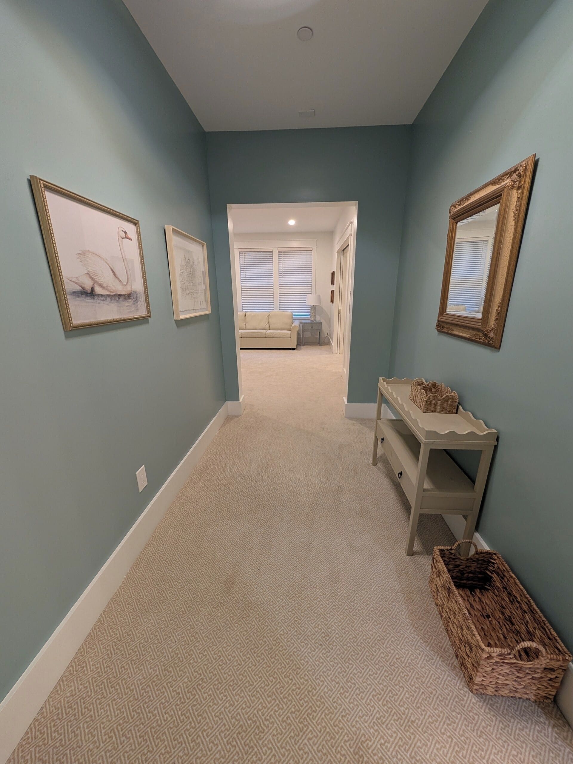 A hallway with light blue walls, a beige carpet, and framed artwork on the walls. There is a bench with baskets and a door leading to a room with a bed and a window.
