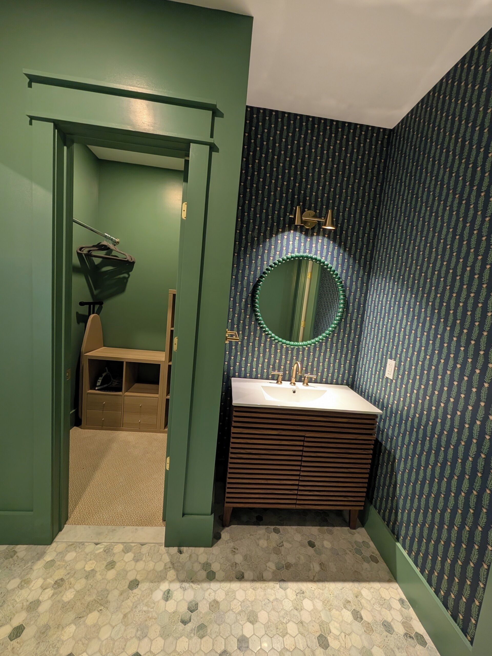 Une salle de bain avec un mur vert, un meuble-lavabo en bois, un miroir rond et un papier peint à motifs.