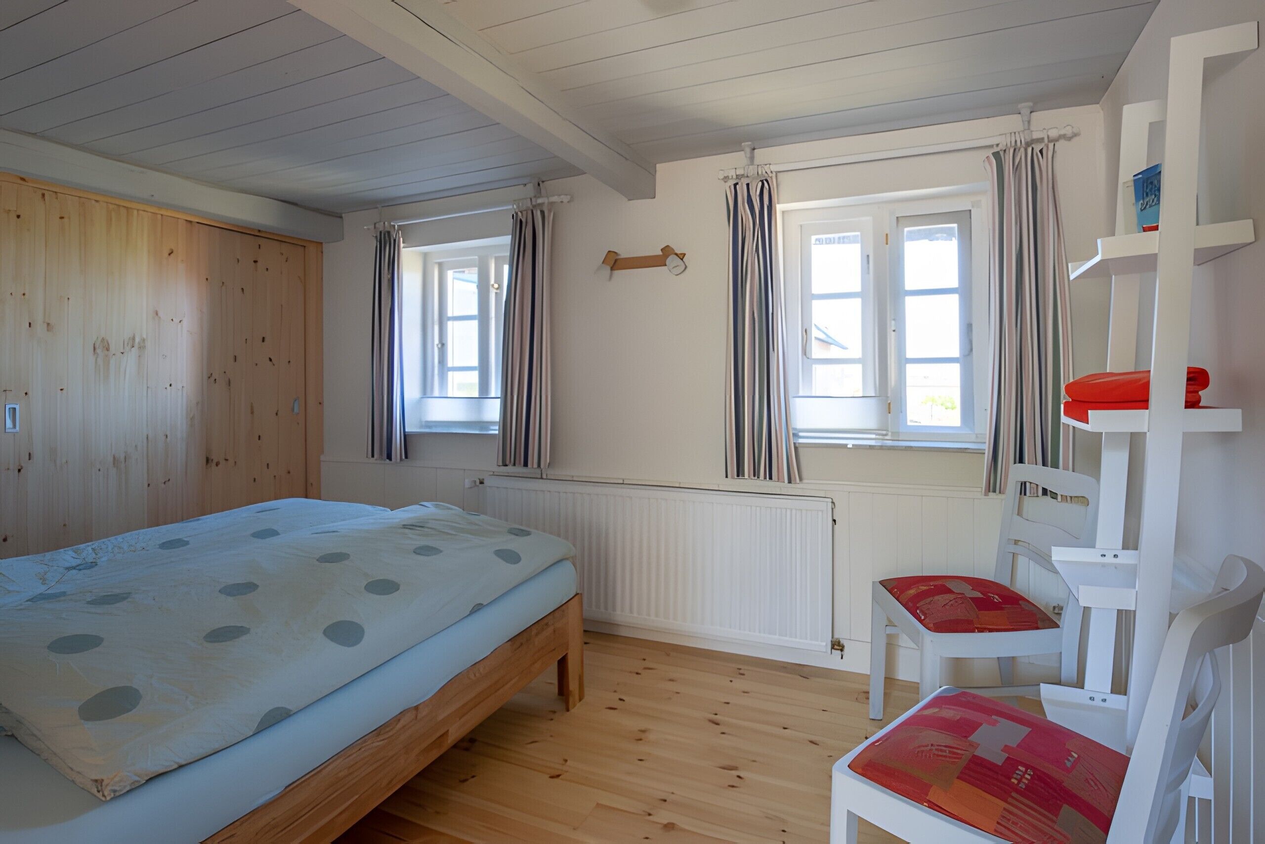 A bedroom with a wooden bed, two windows with curtains, a radiator, and a white chair with a red cushion.