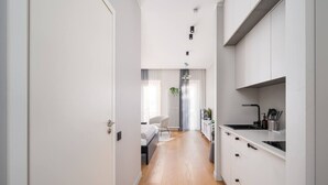 Ein modernes, minimalistisches Hotelzimmer mit Holzboden, einem Bett mit weißen Leinentüchern, einer kleinen Sitzecke mit einem Sessel und einer Pflanze sowie einer Küchenzeile mit weißen Schränken und einer schwarzen Arbeitsplatte.