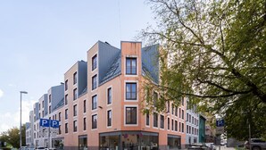 Ein mehrstöckiges Gebäude mit modernem architektonischen Design, das Glas- und Metallfassaden kombiniert, und mit mehreren Parkplätzen, die durch blaue Schilder gekennzeichnet sind.