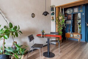 A dining area with a round table, chairs, and a red table runner. There are potted plants and a hanging plant in the room. - Dark & Moody Mid Century Apartment (Riga)