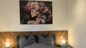 A bedroom with a bed, two bedside tables with lamps, a headboard, and a large floral artwork on the wall. - House 2: 8 high-quality vacation homes with private sauna & jacuzzi on Sweet Lake (Seegebiet Mansfelder Land)