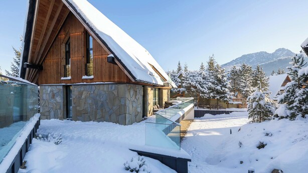 Un edificio moderno de madera con paredes de piedra, barandilla de cristal y un paisaje nevado con montañas al fondo.