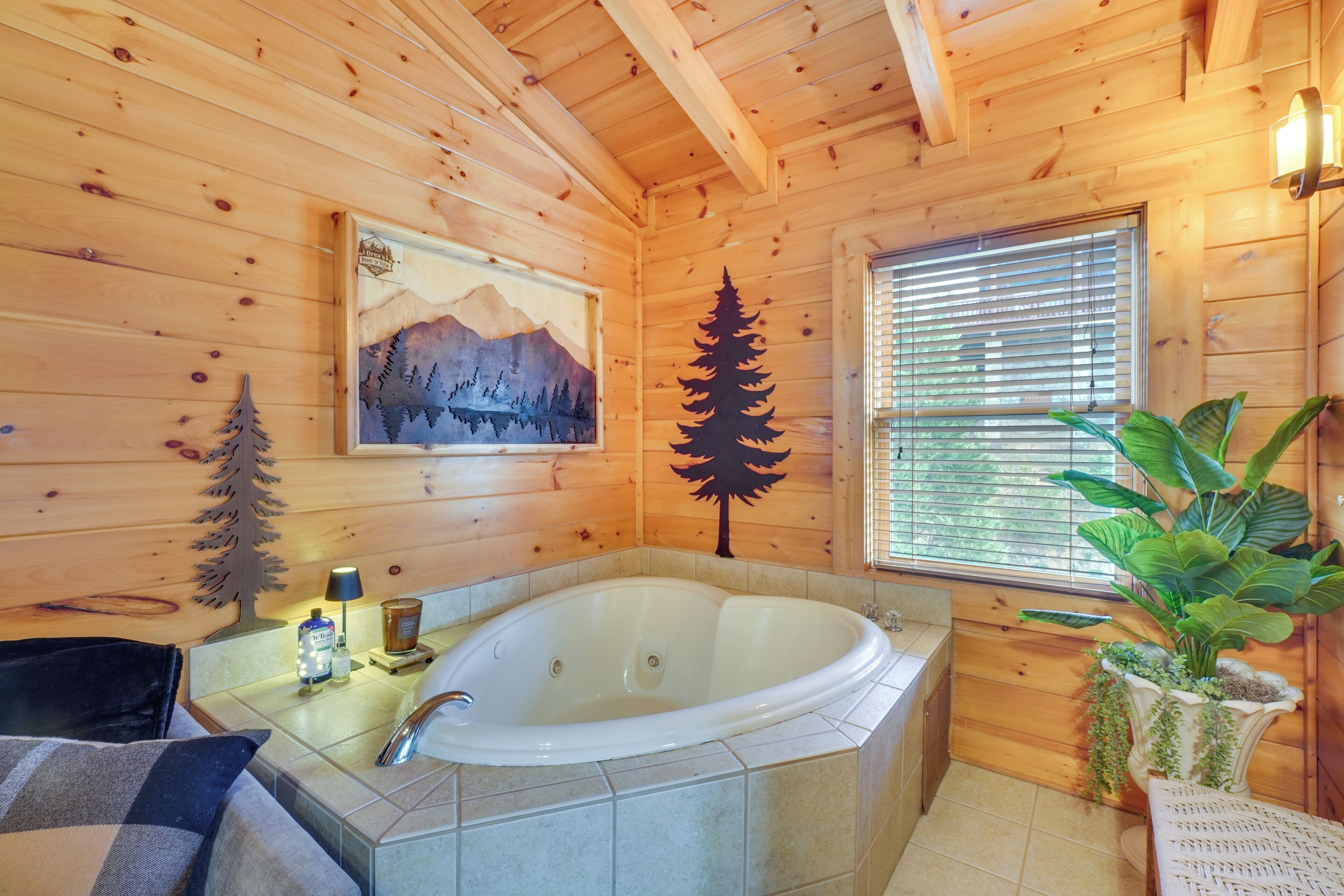 A wooden cabin bathroom with a large bathtub, a window with blinds, and a painting of mountains.