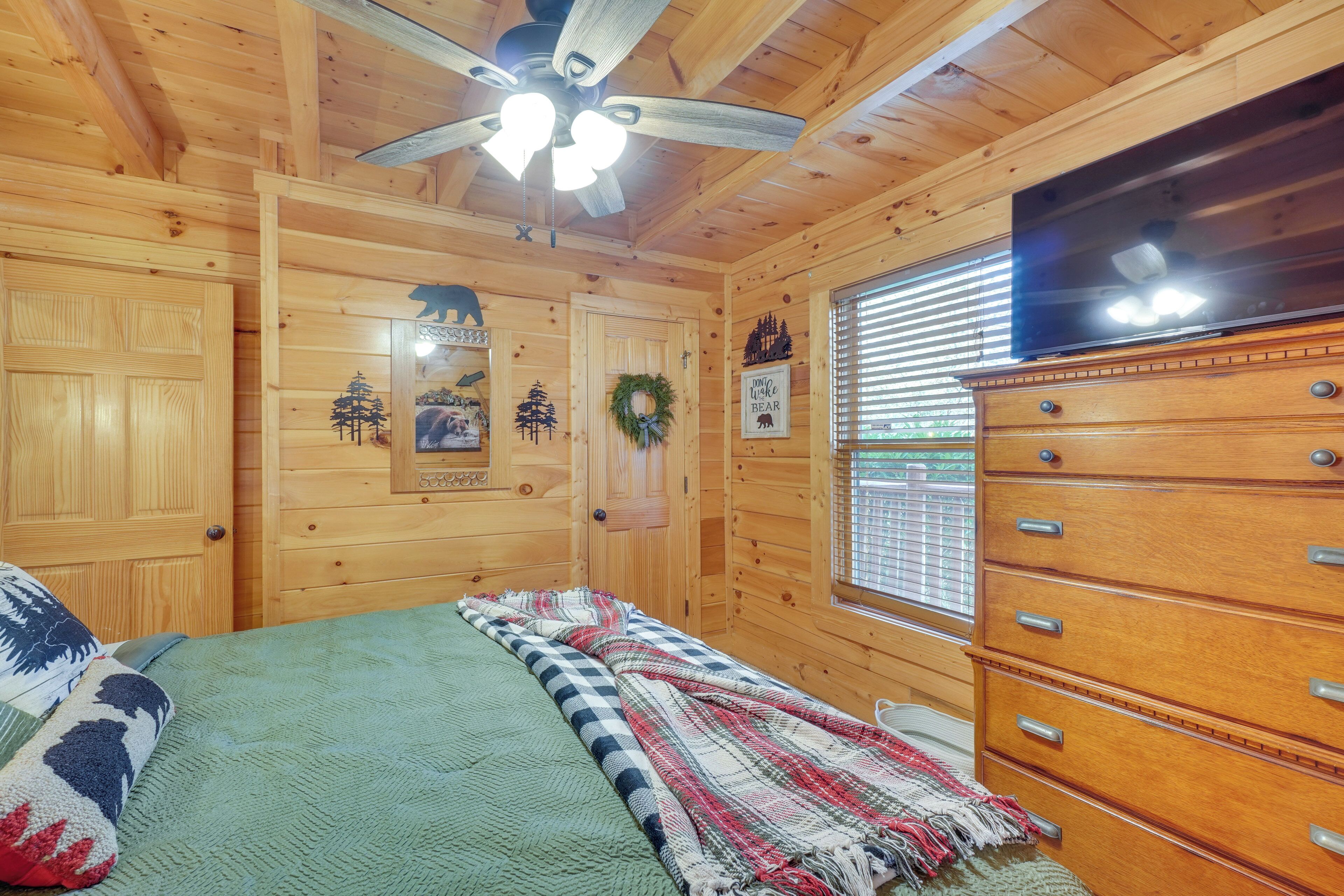 A wooden cabin room with a bed, a dresser, a television, and a ceiling fan.