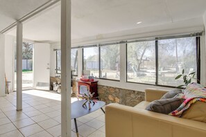 A spacious living area with a large window, a beige sofa, a small table, and a stone wall. - 18 Mi to Joshua Tree NP: Wfh-ready Desert Escape (Morongo Valley)