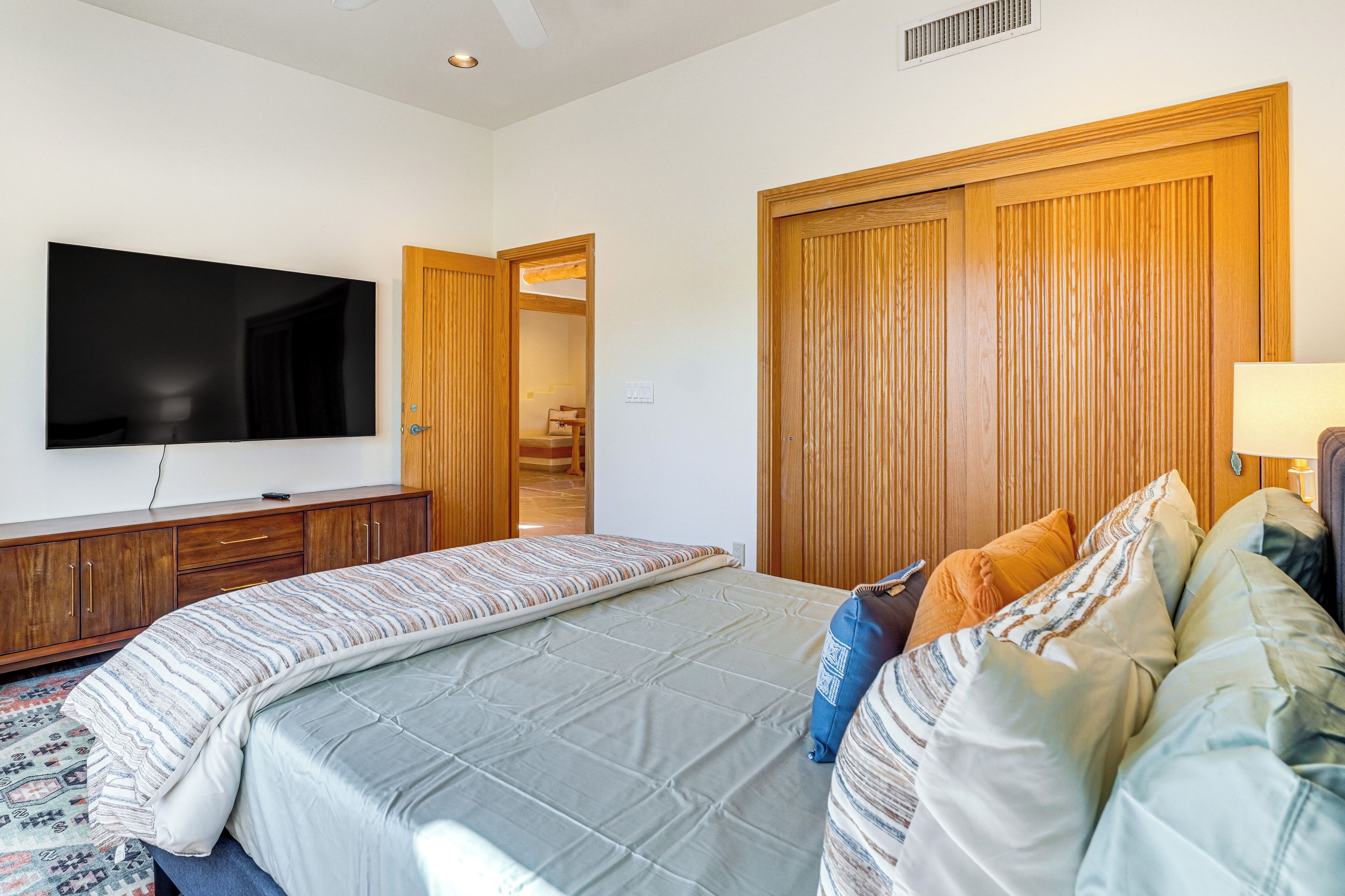 A bedroom with a large bed, a television mounted on the wall, wooden wardrobes, and a door leading to another room.