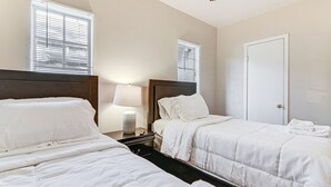 Ein Hotelzimmer mit zwei Betten, einem Nachttisch mit Lampe, einer Tür und Fenstern mit Jalousien.