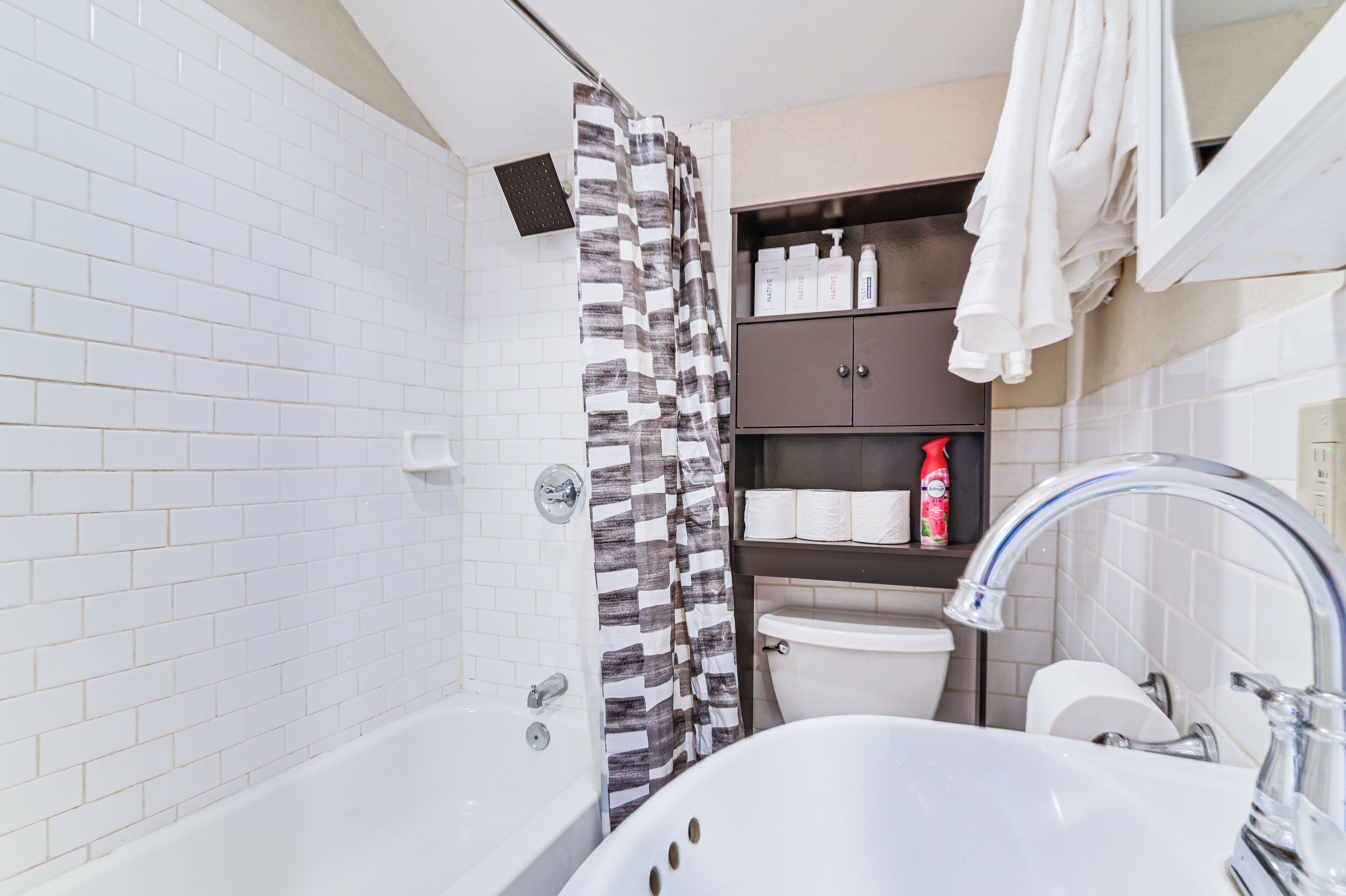 A bathroom with a white tiled wall, a shower with a patterned curtain, a toilet, a sink with a faucet, and shelves with toiletries.