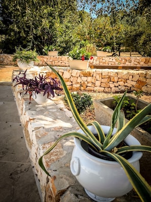 Un muro di pietra con piante in vaso, una panchina in pietra e un’area giardino con diverse piante e alberi.