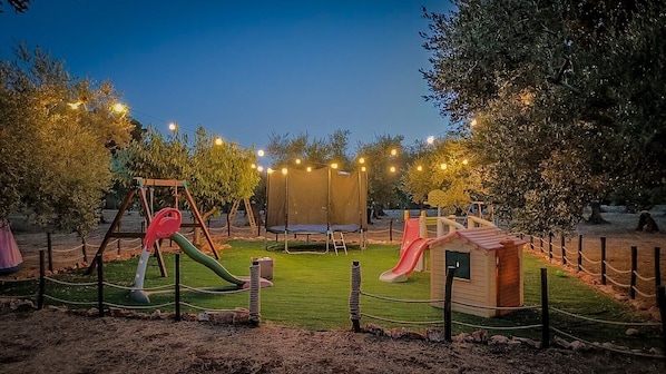 Un'area giochi con altalene, uno scivolo e una piccola casetta, circondata da alberi e luci decorative.
