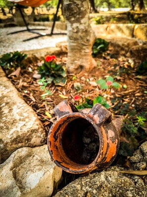 Un’ambientazione esterna rustica con un vaso di terracotta consumato, un albero e un’aiuola piena di piante.