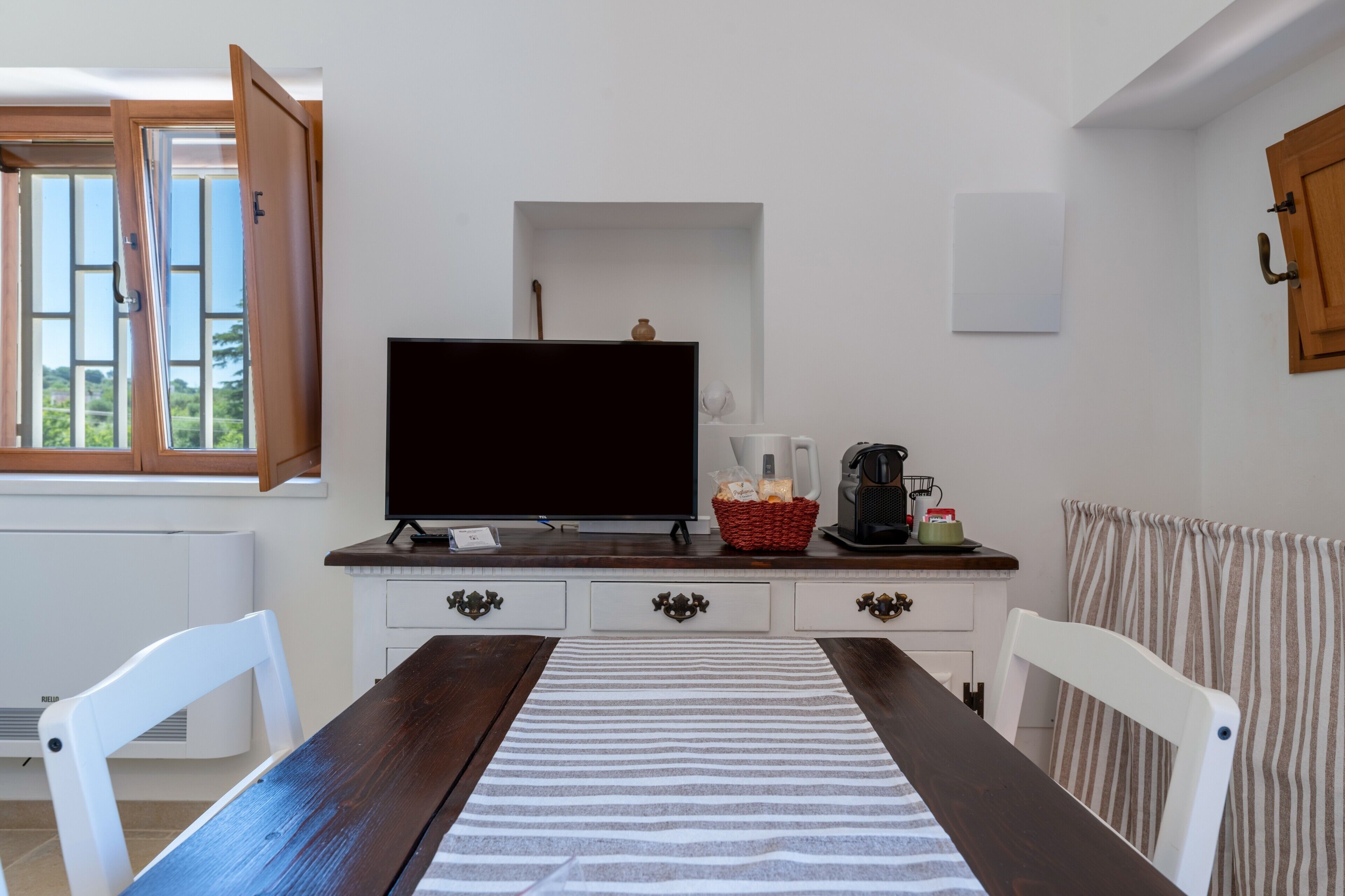 Una habitación con una mesa de madera, sillas blancas, un televisor, una cesta con aperitivos, una cafetera y una ventana con contraventanas abiertas.