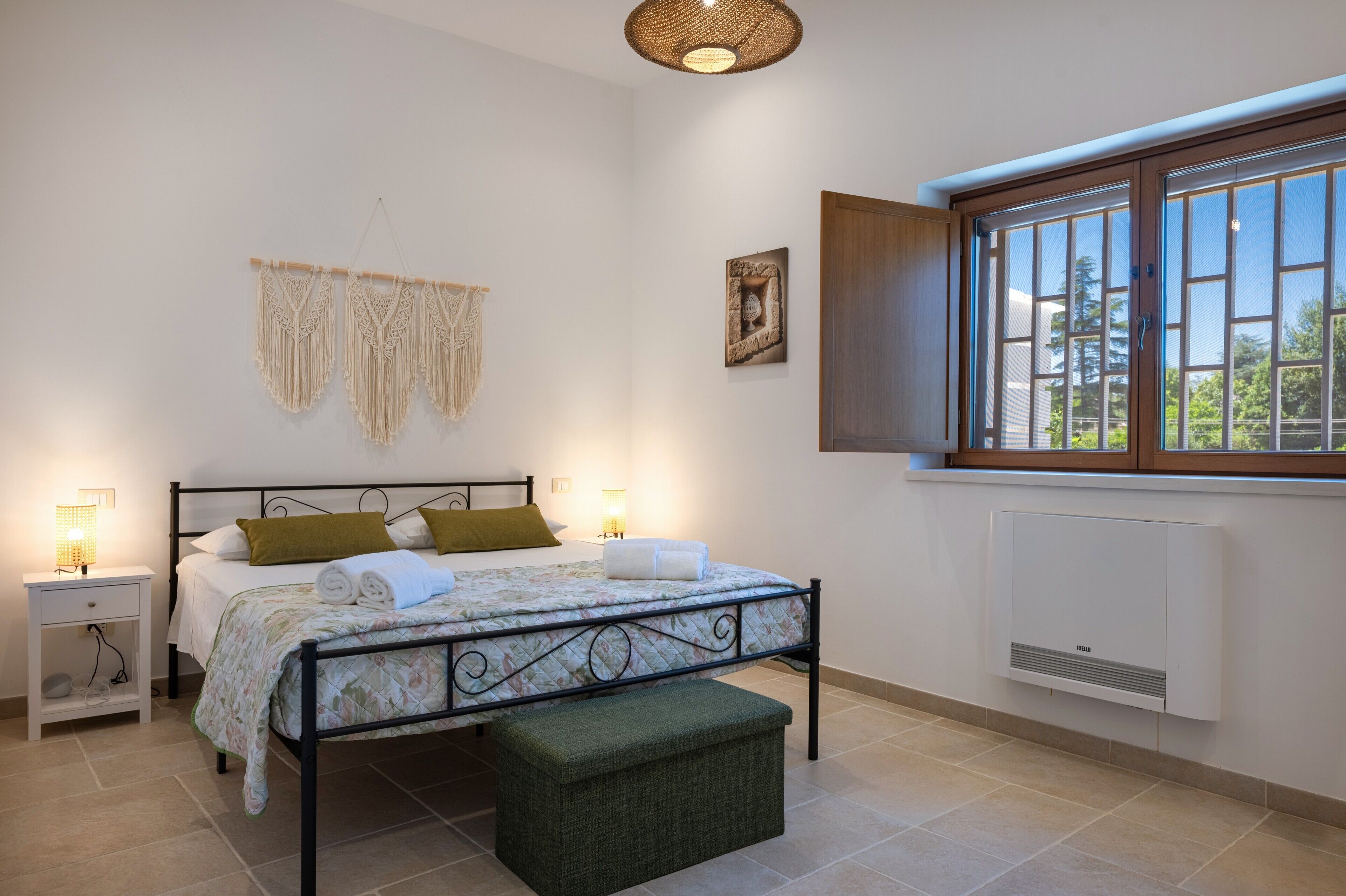 Een slaapkamer met een bed, nachtlampjes, een raam, een wandairconditioner en macramé wanddecoraties.