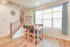 A dining area with a wooden table and chairs, a clock on the wall, and a window with curtains. - Creekside Retreat Near Olde Town Arvada (Arvada)