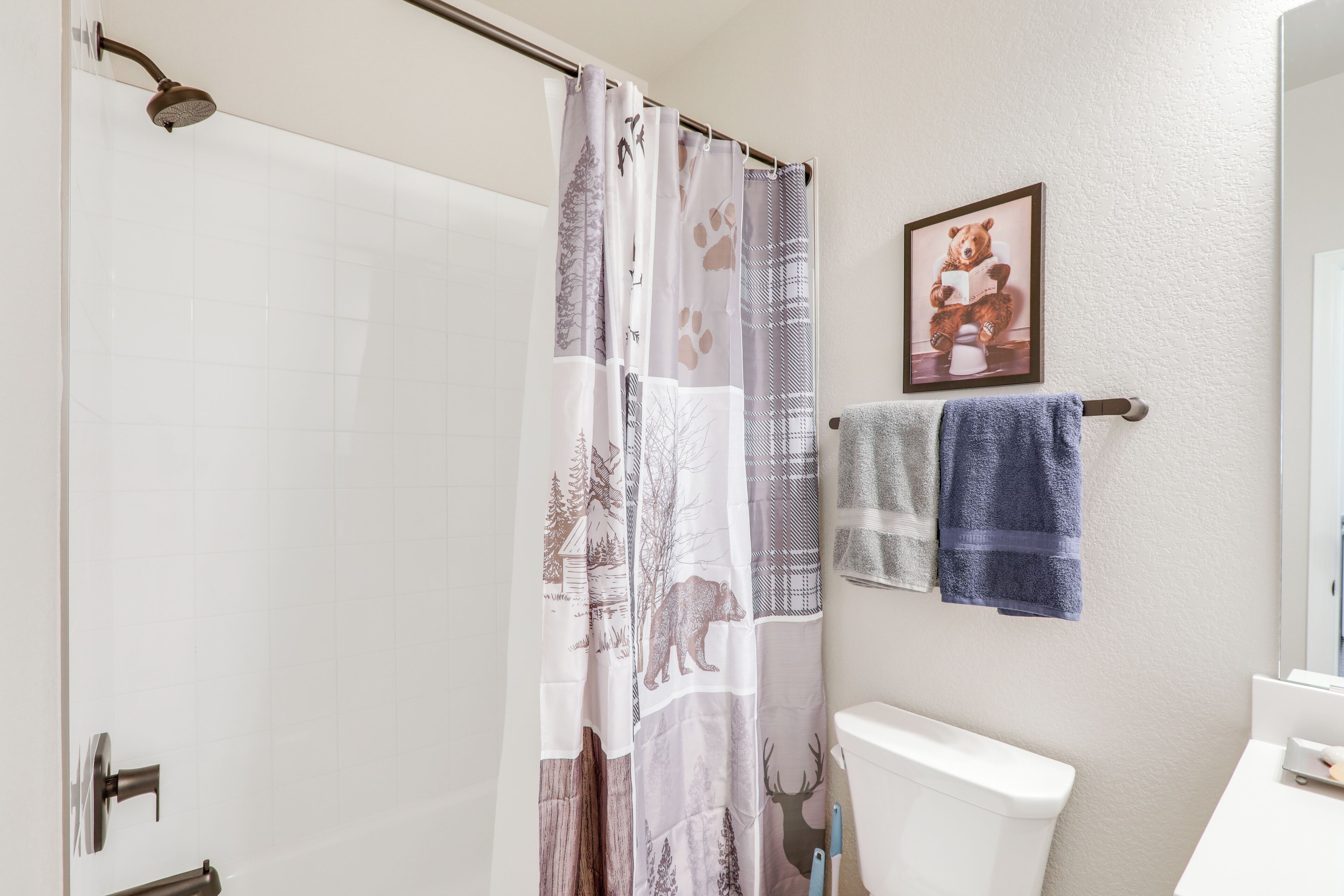 A bathroom with a shower curtain featuring forest and animal prints, a toilet, a towel rack with two towels, and a framed picture on the wall.