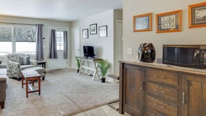 Un salón con sofá, sillón, mesa de centro, televisor y escritorio con computadora. Se observan cuadros en la pared y un microondas en la encimera.