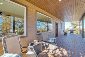 A wooden deck with outdoor seating, a grill, and large windows offering views of trees and a clear sky.