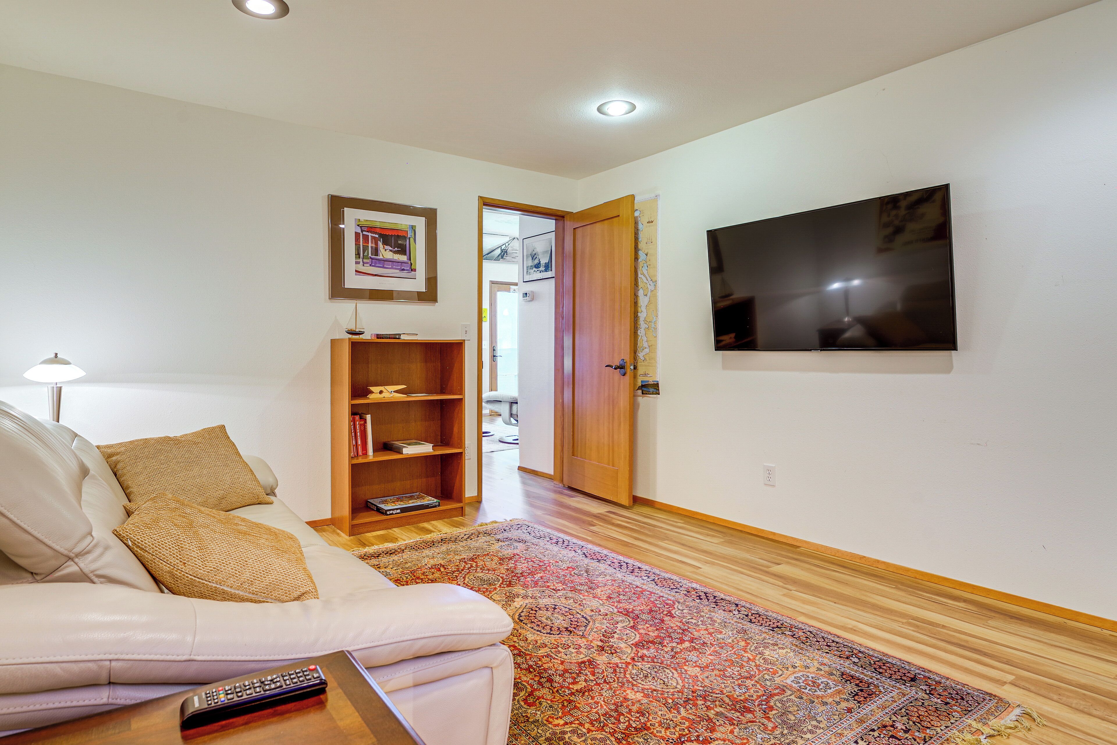 Ein Wohnzimmer mit einer Couch, einem Couchtisch mit Fernbedienung, einem Holzregal, einem gemusterten Teppich und einem an der Wand montierten Fernseher.