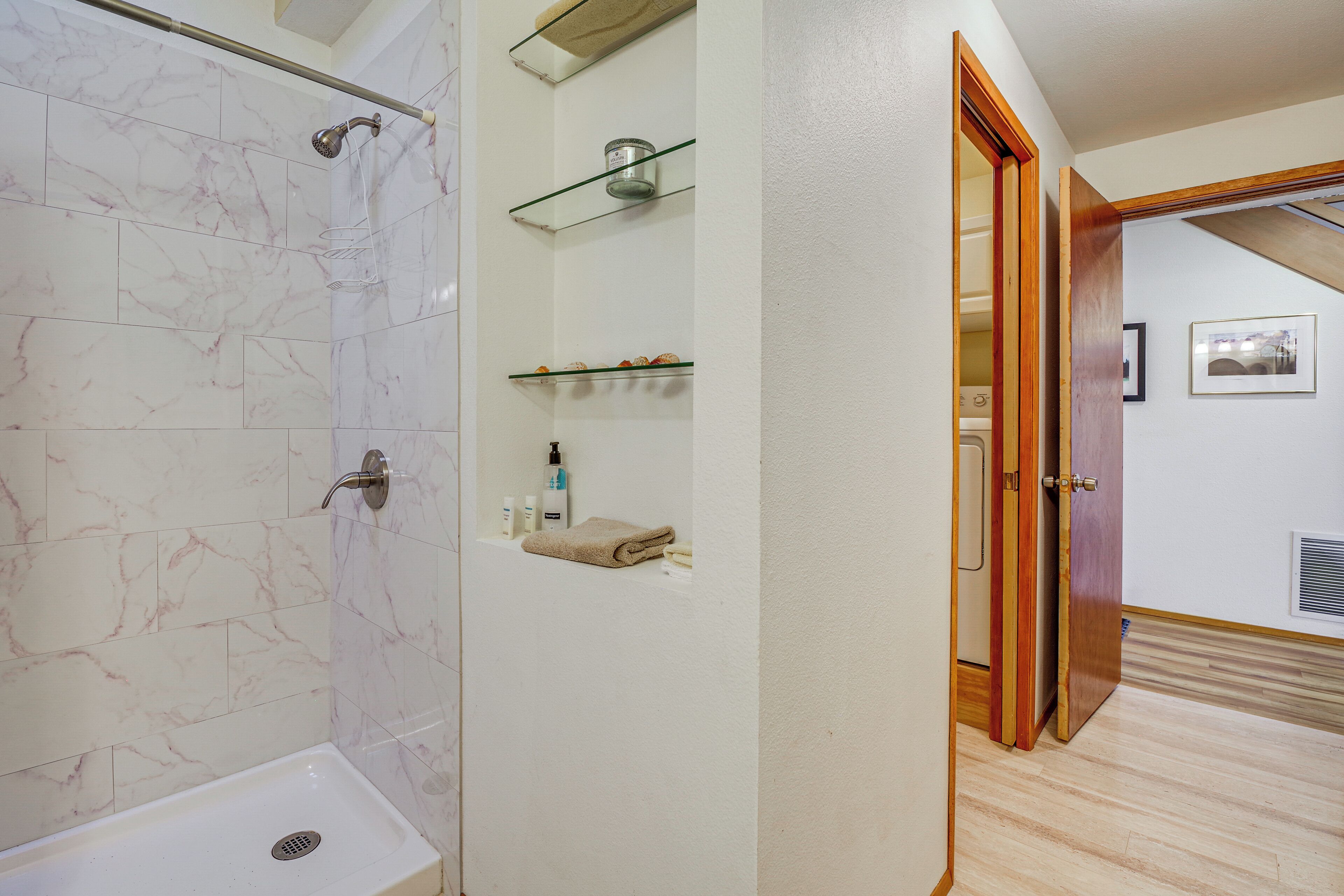 A bathroom with a shower, a sink, marble tiles, glass shelves with toiletries, and a wooden door leading to another room.