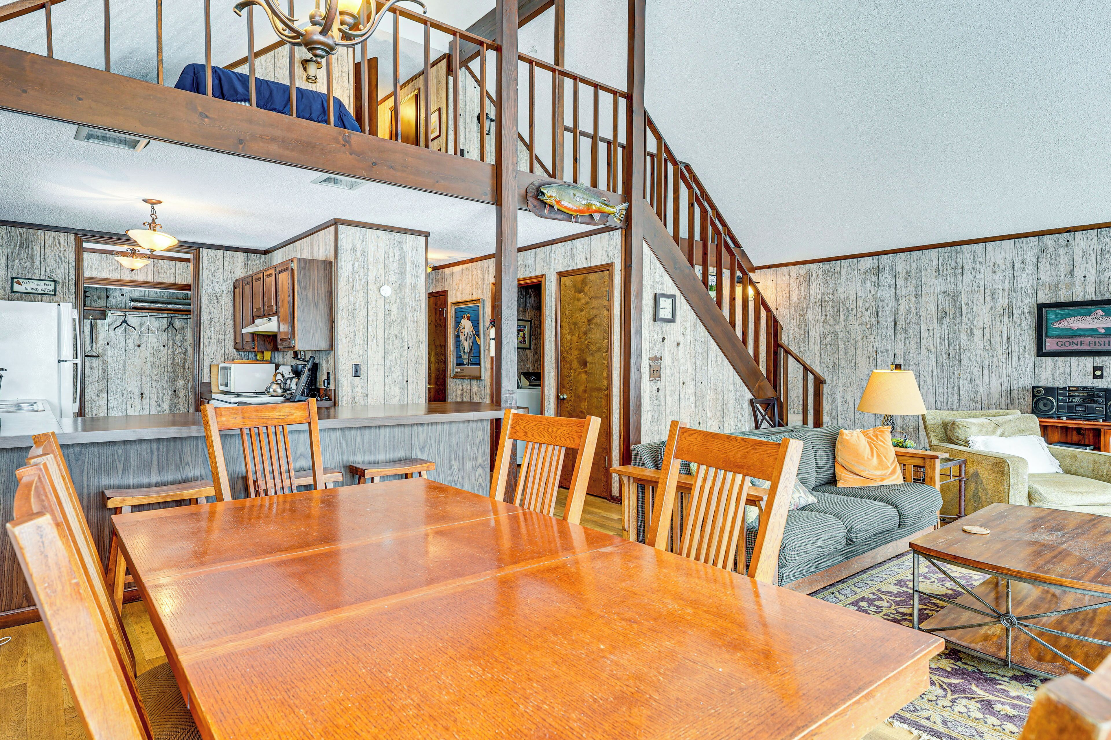 A spacious living area with a large wooden dining table, a staircase, and a kitchen in the background.