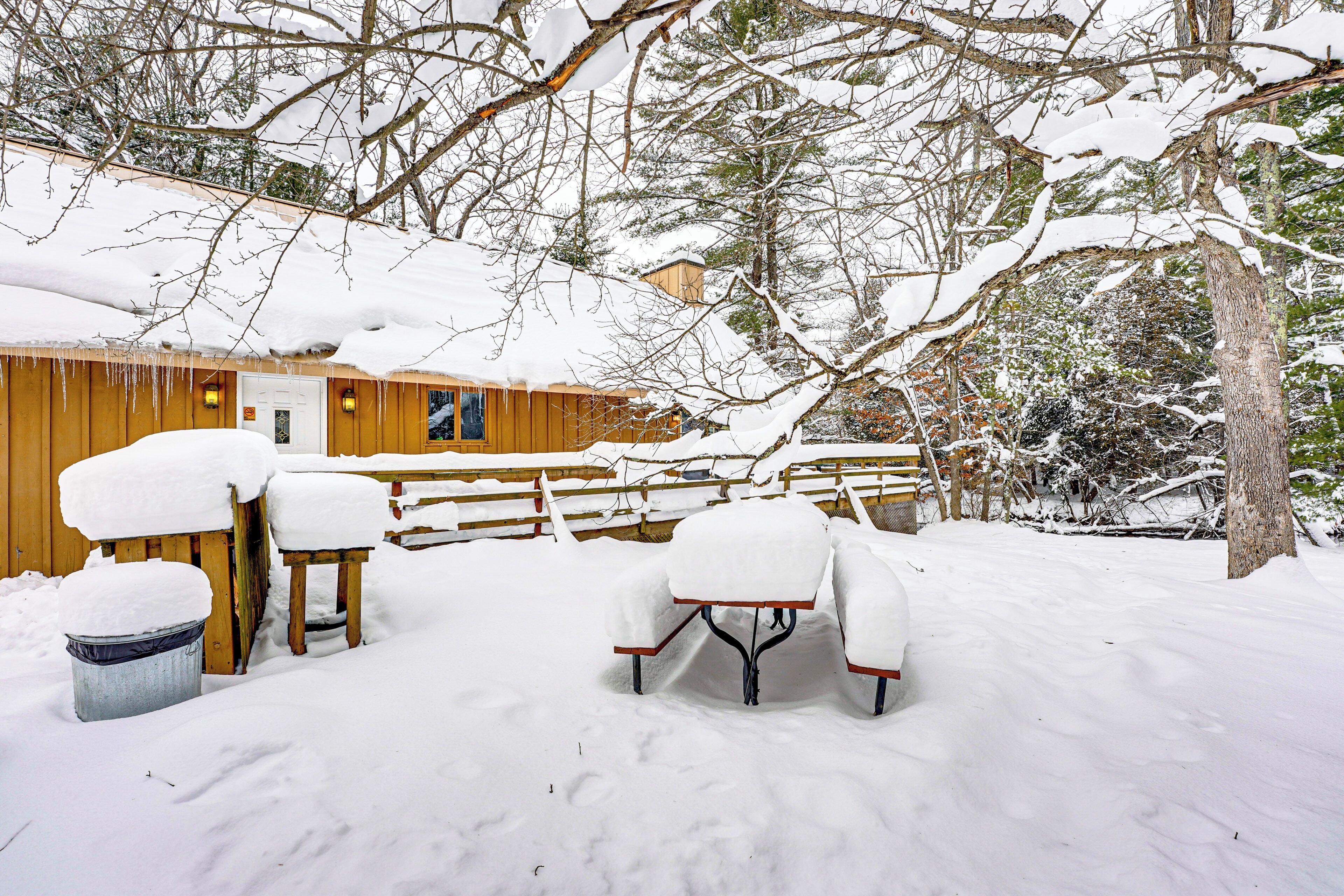 A wooden cabin with snow-covered tables and chairs, a wooden fence, and trees in the background.