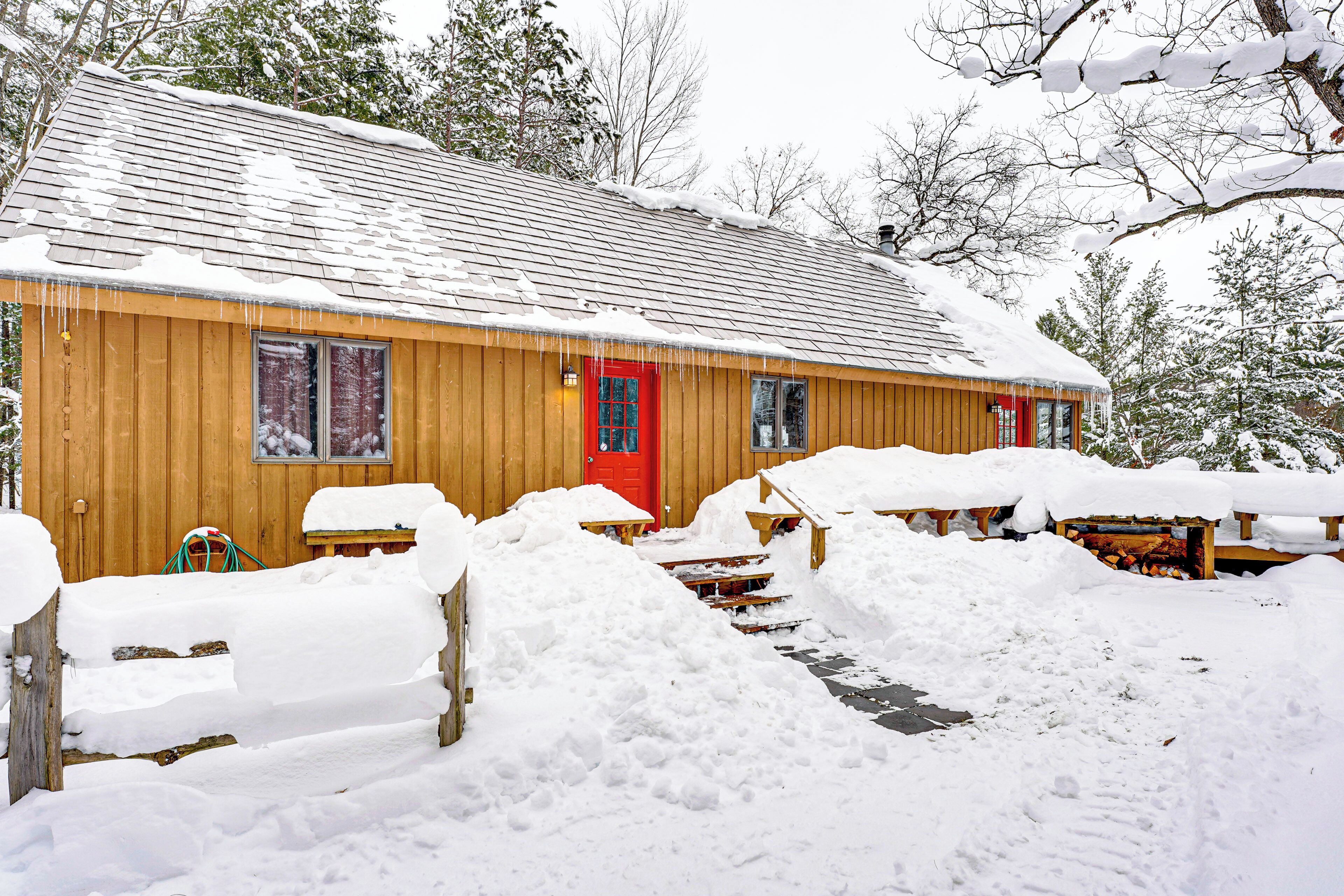 Una cabaña de madera con una puerta roja, rodeada de nieve y árboles.
