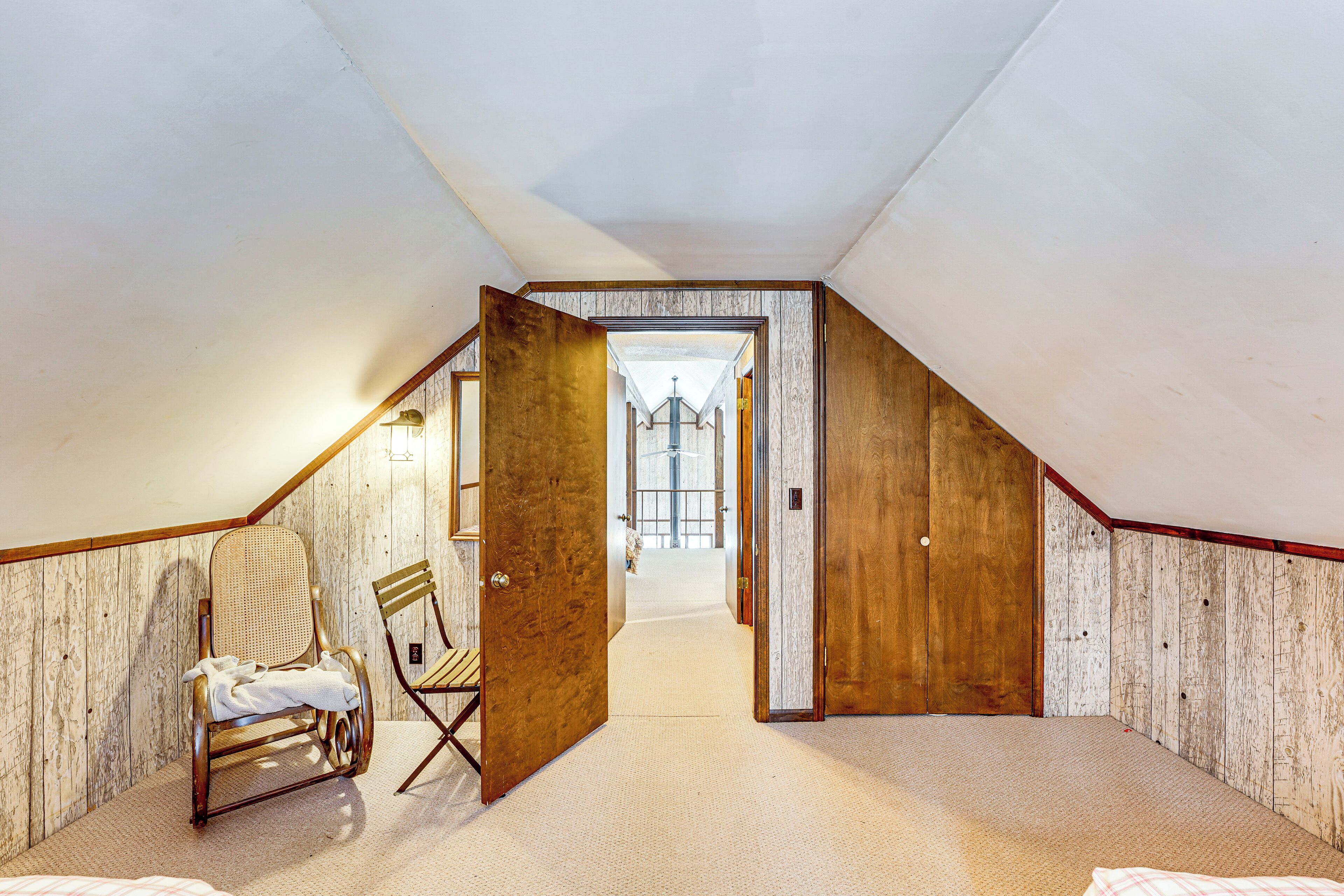Een zolderkamer met schuine wanden, houten lambrisering, een groot raam en een stoel.