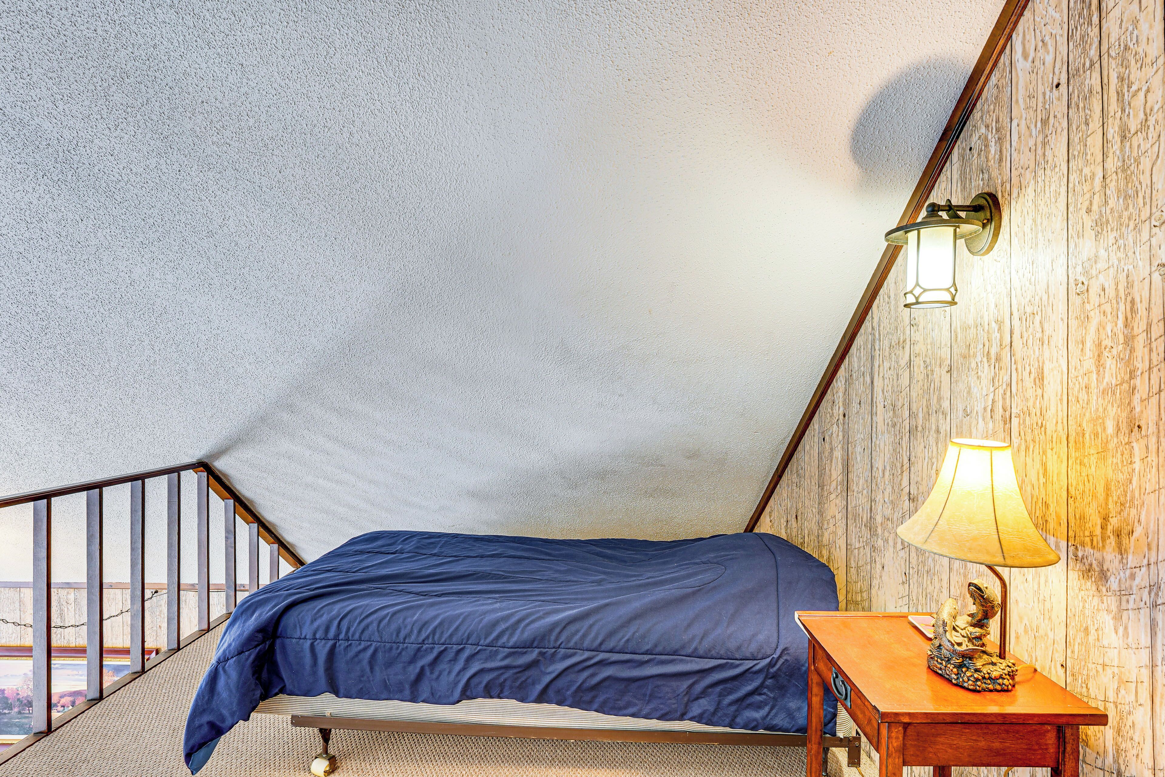A bedroom with a sloped ceiling, a bed with a blue cover, a wooden nightstand with a lamp, and a view of the outdoors.