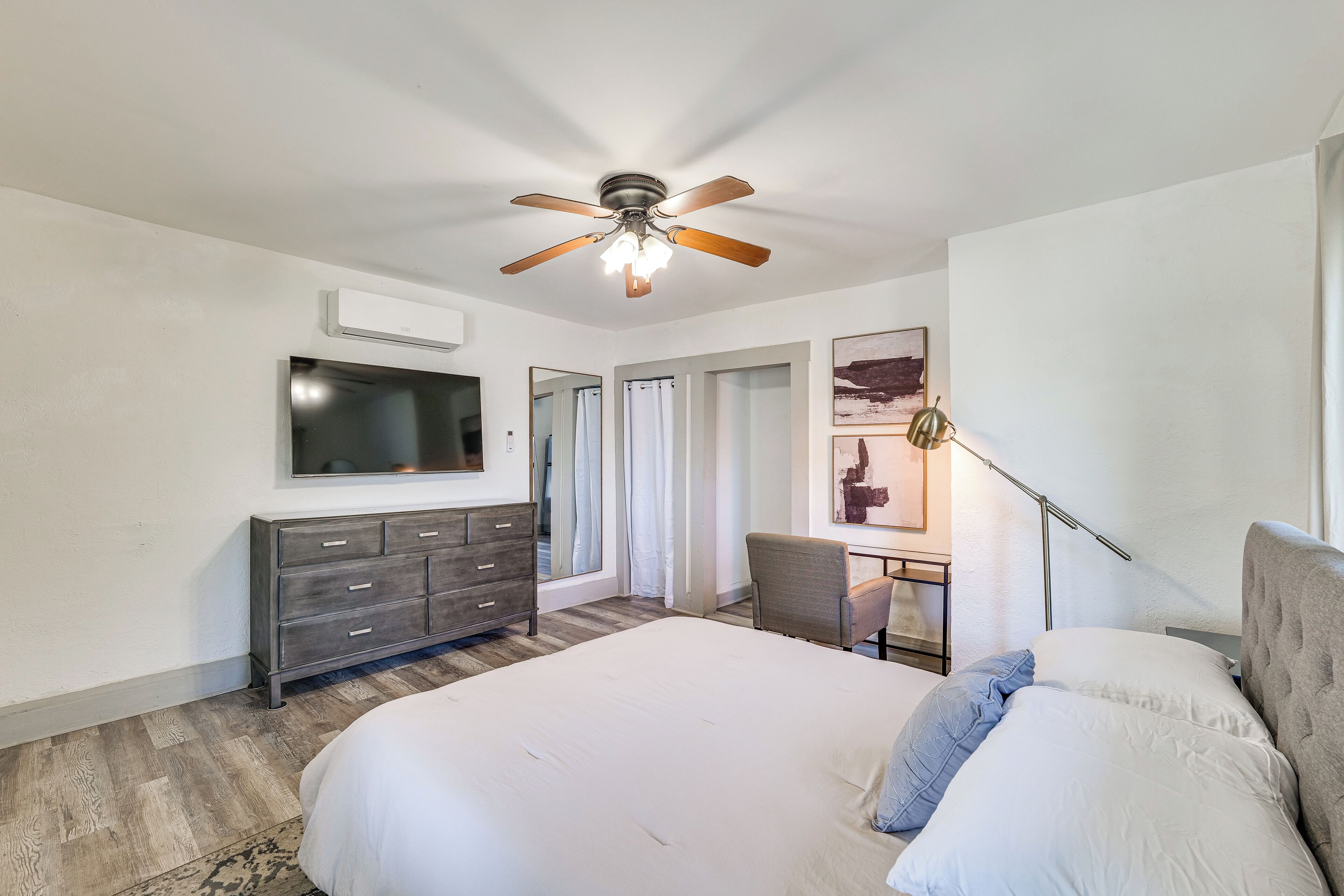A bedroom with a large bed, a ceiling fan, a TV, a dresser, and a desk with a lamp.