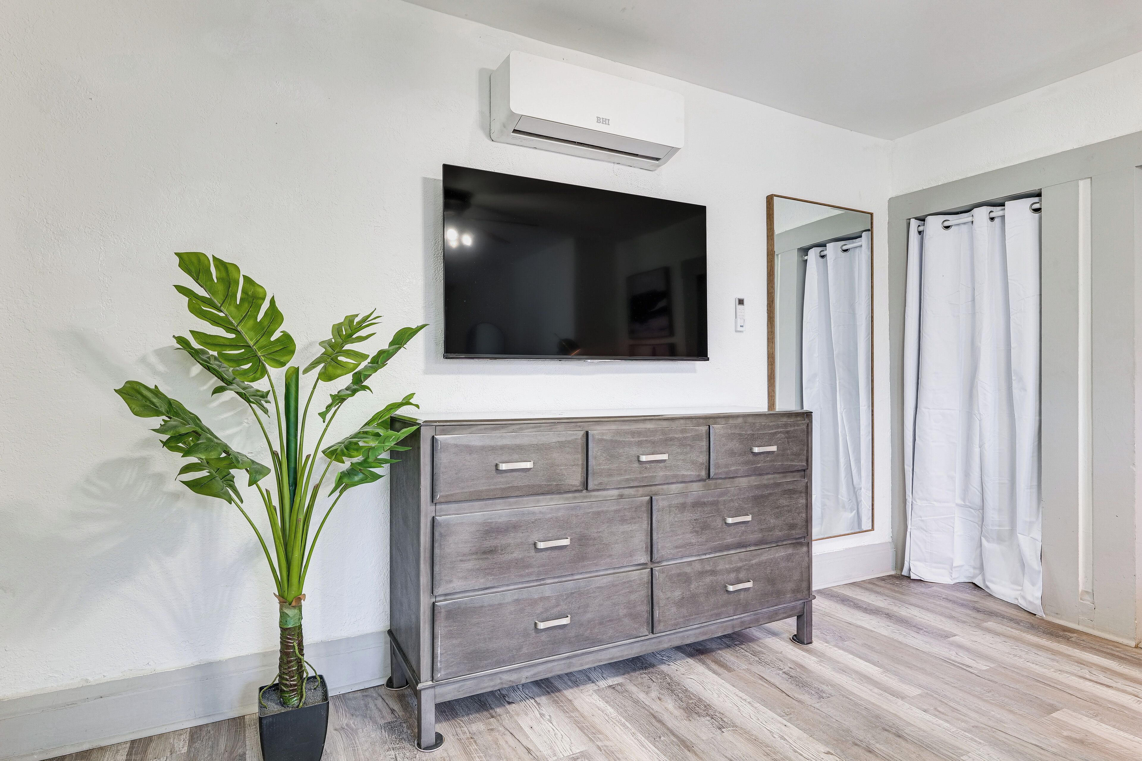 A modern room with a grey dresser, a large flat-screen TV mounted above it, a potted plant, and a shower area with white curtains.