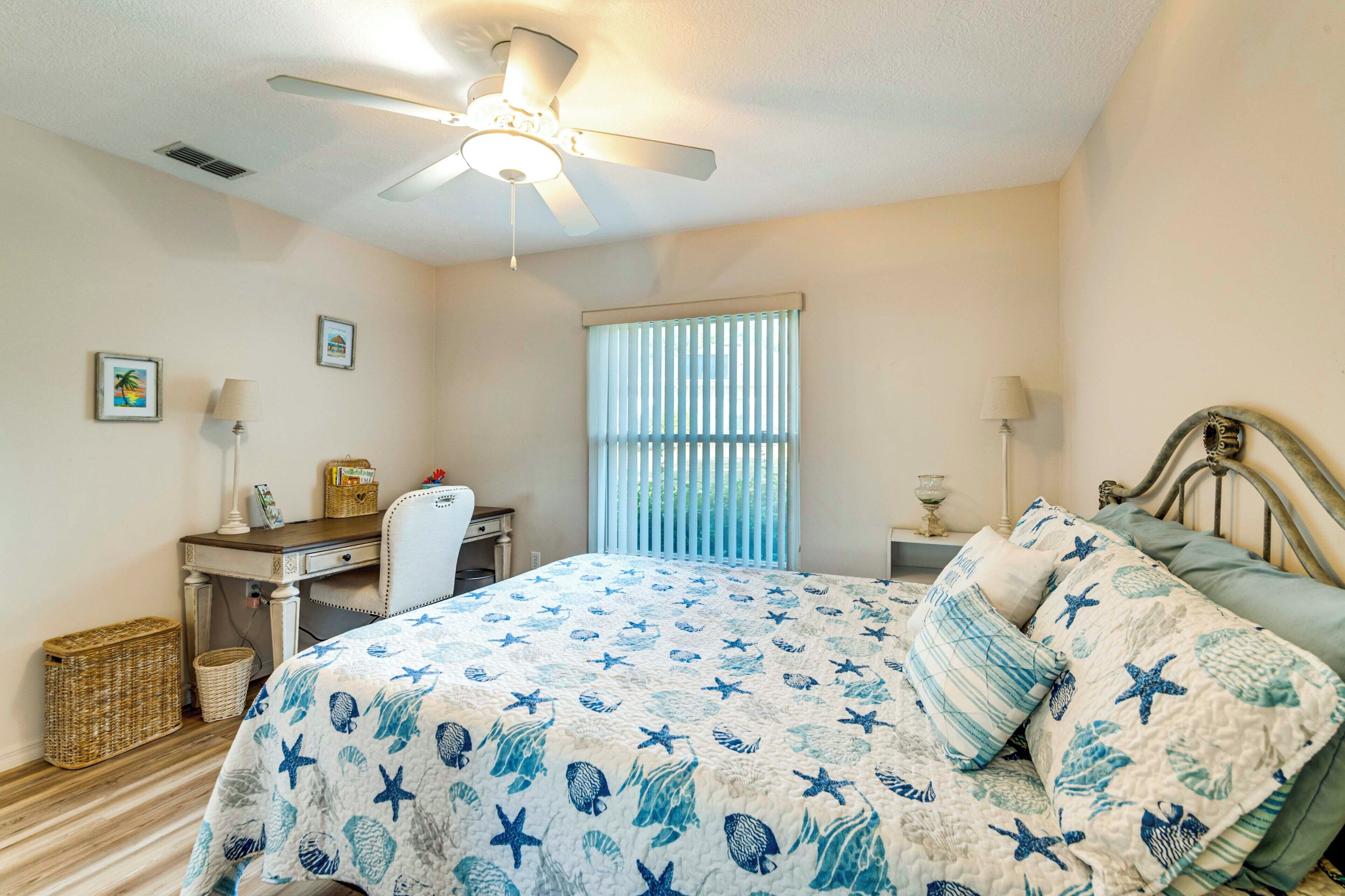 A bedroom with a bed, a desk, a chair, a ceiling fan, and a window with blinds.