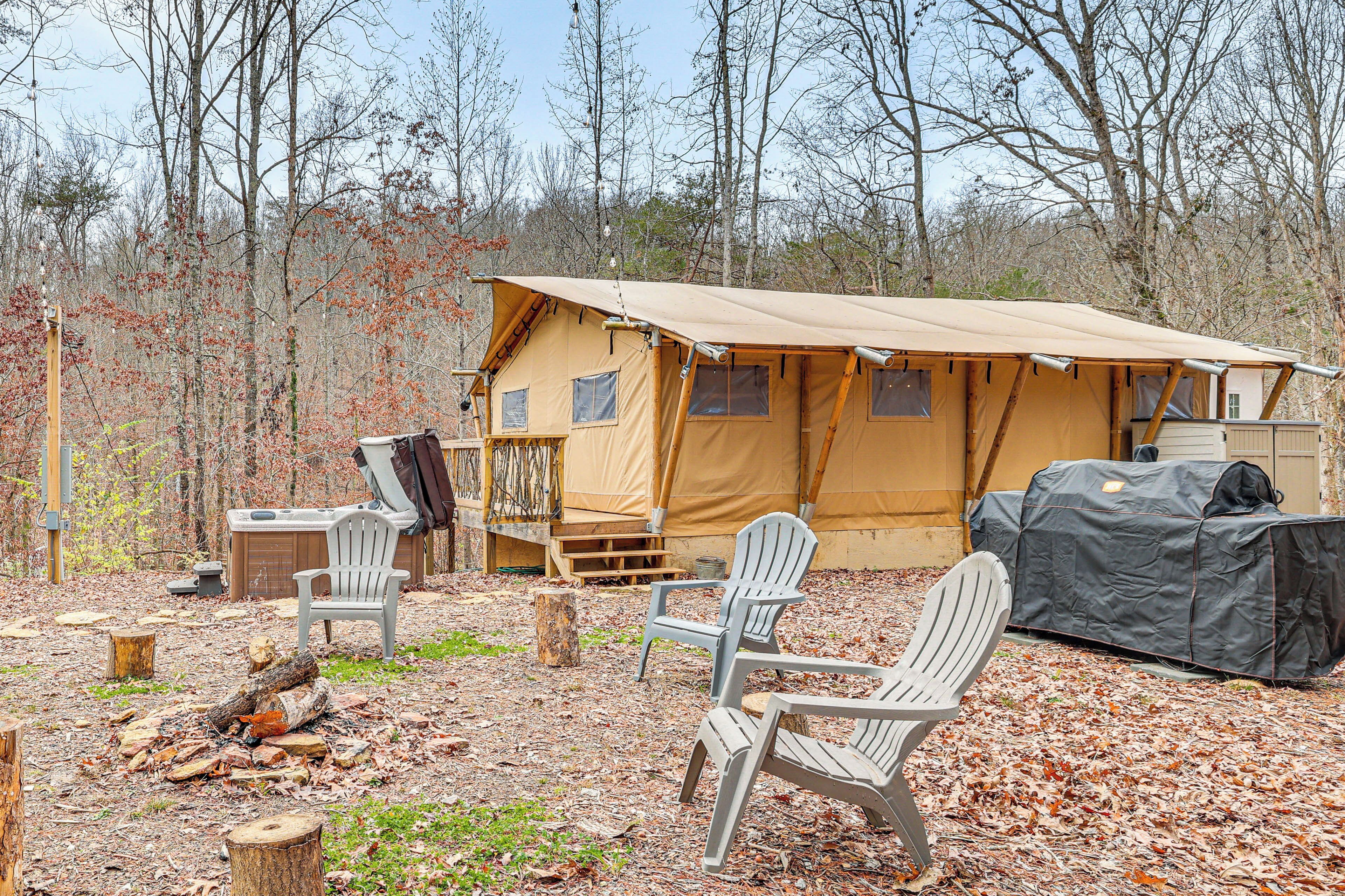 A tented campsite with a covered grill, two Adirondack chairs, and a fire pit.