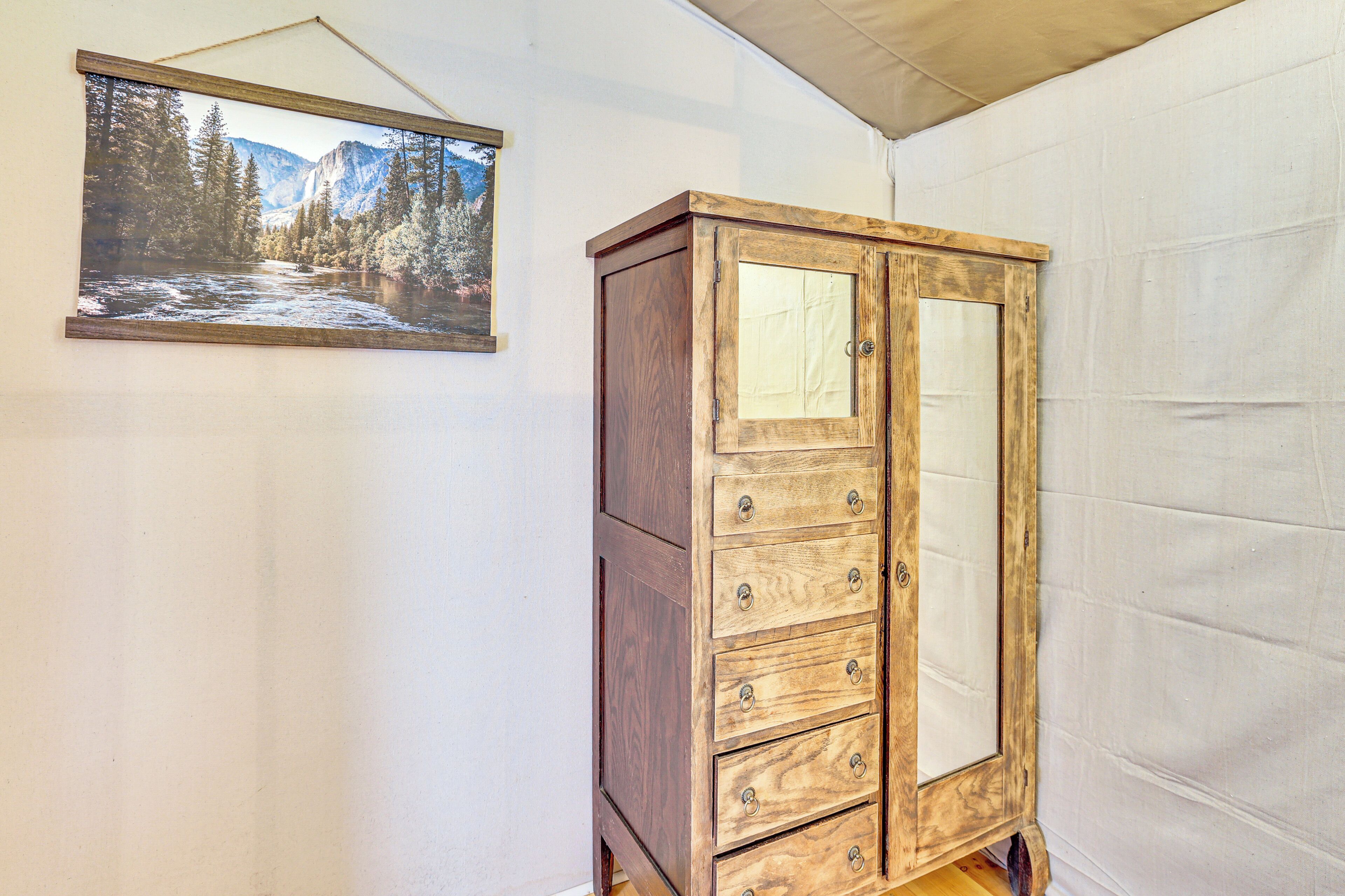 A wooden cabinet with drawers and a glass door, a framed landscape painting on the wall, and a white wall.